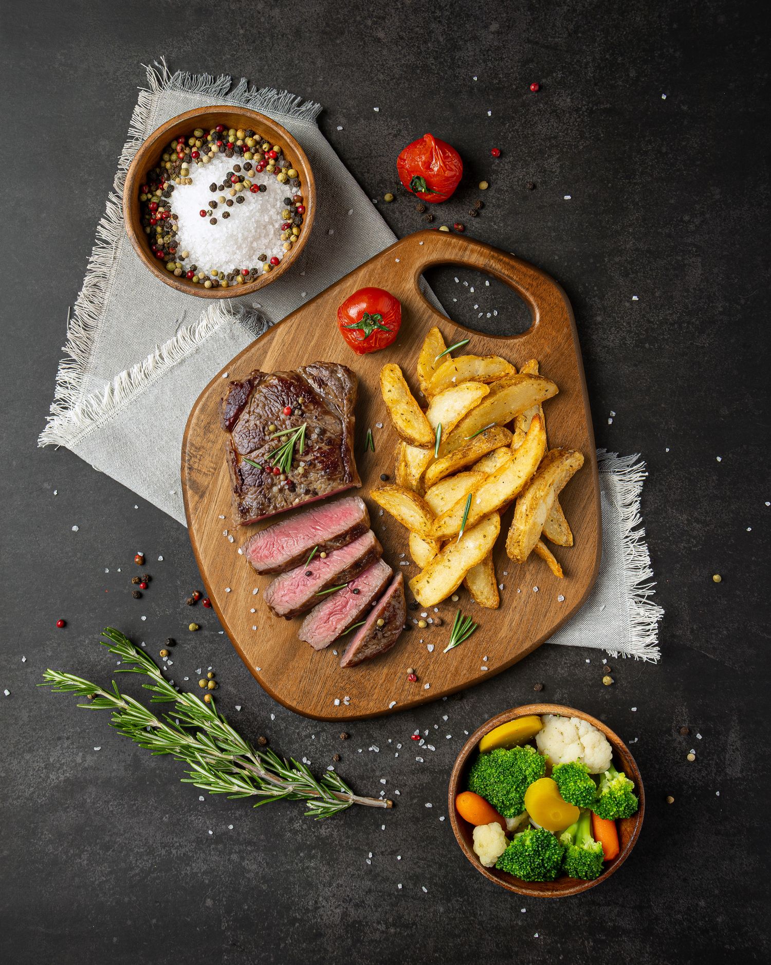 Juicy grilled steak with golden potato wedges and fresh herbs served on a rustic wooden board with seasoning.
