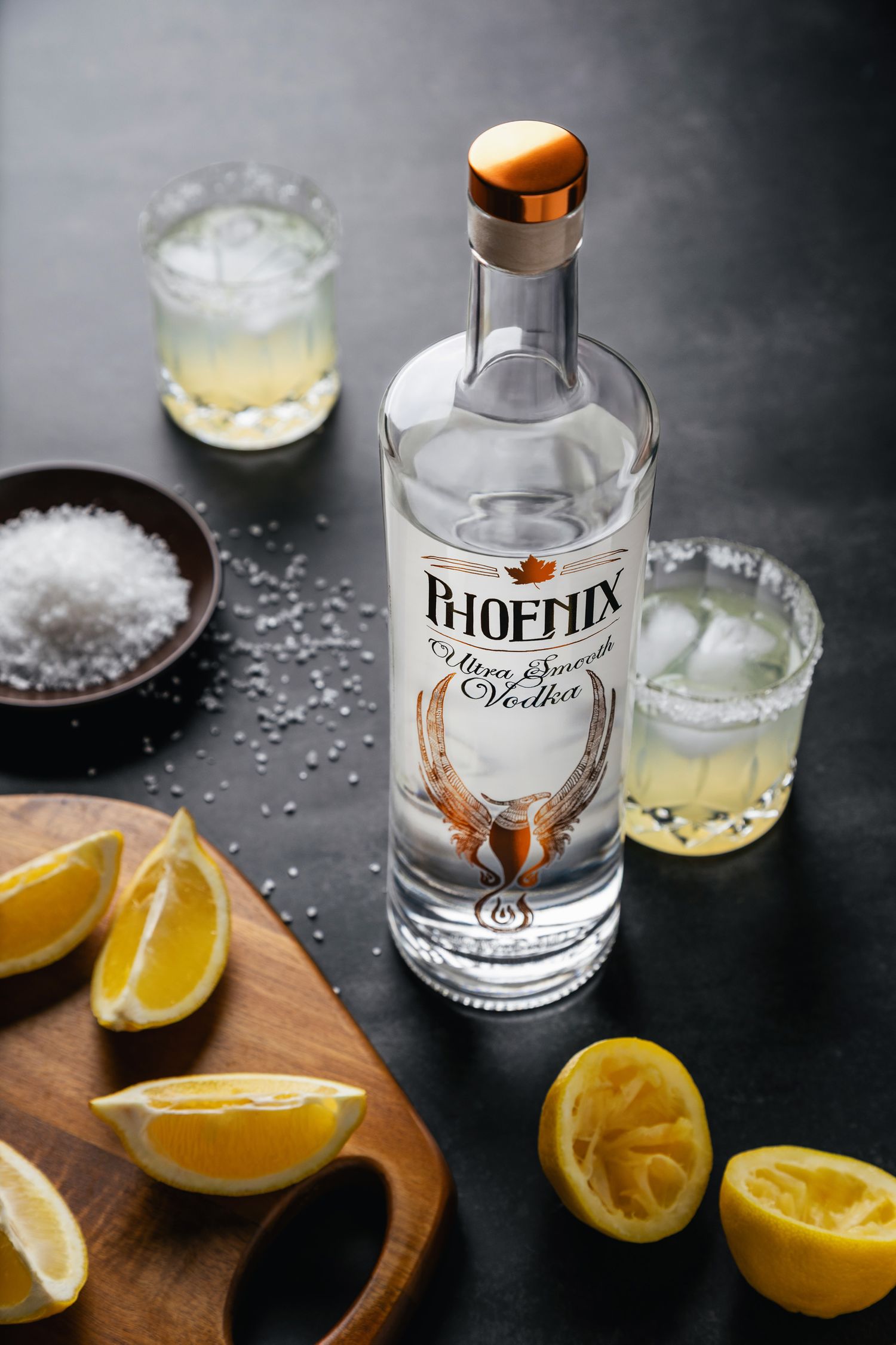 Phoenix Ultra Smooth Vodka bottle with lemon wedges, salted rim glasses, and coarse salt on dark background.