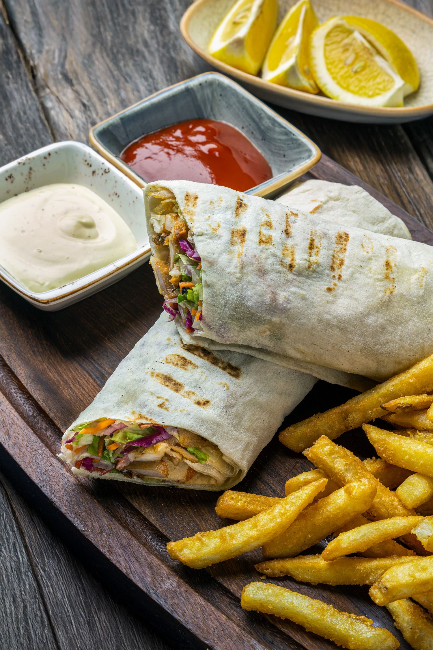 Chicken wrap sandwich served with crispy seasoned french fries and dipping sauces on a rustic wooden table.