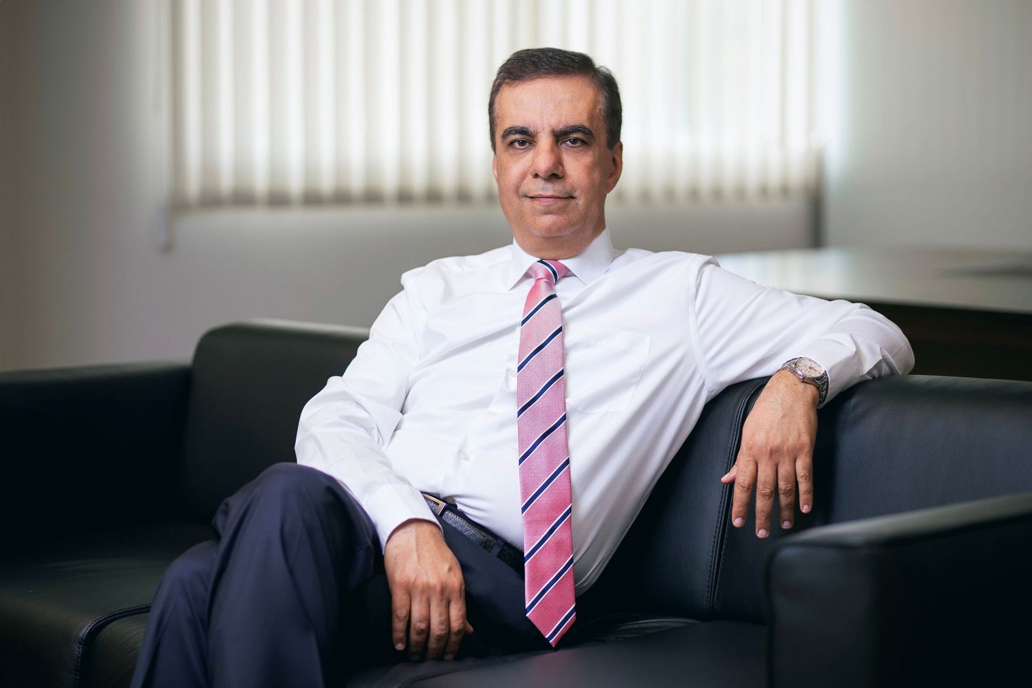 Professional businessman in white shirt and pink striped tie sitting relaxed on a dark sofa in a modern office.