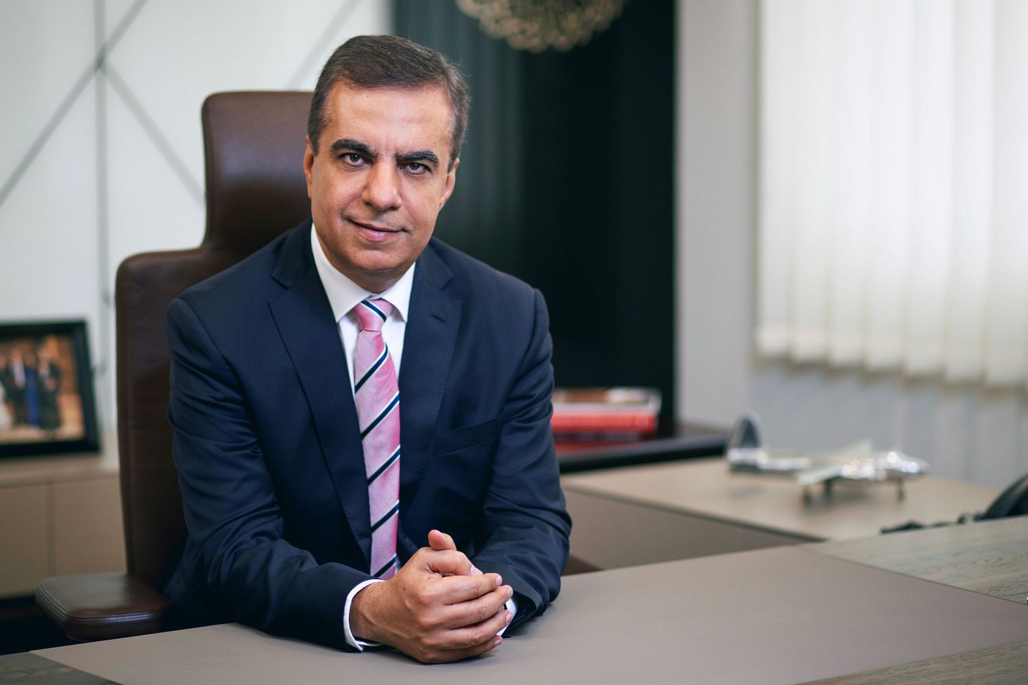 Professional businessman in navy suit sitting at desk in modern office, posing confidently with hands clasped.
