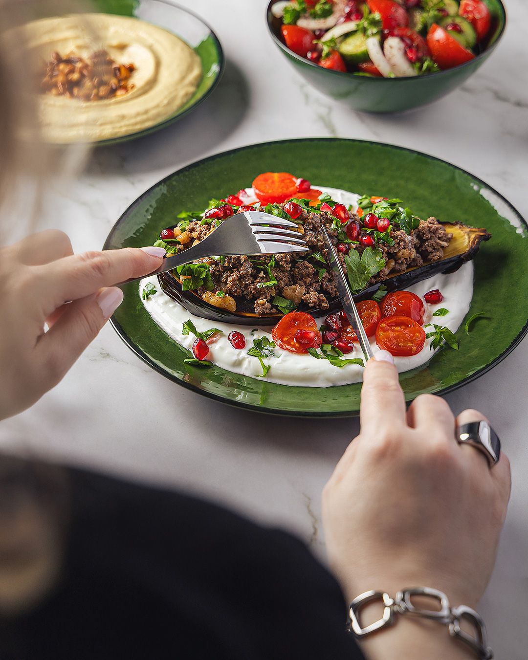 Hands cutting stuffed eggplant with ground meat, tomatoes, pomegranate on white yogurt sauce, green plate.