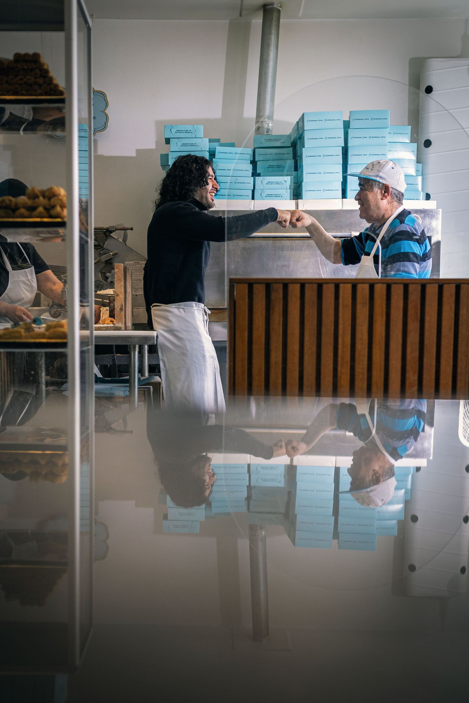 Two bakery workers fist bumping over a counter with light blue boxes stacked in the background.