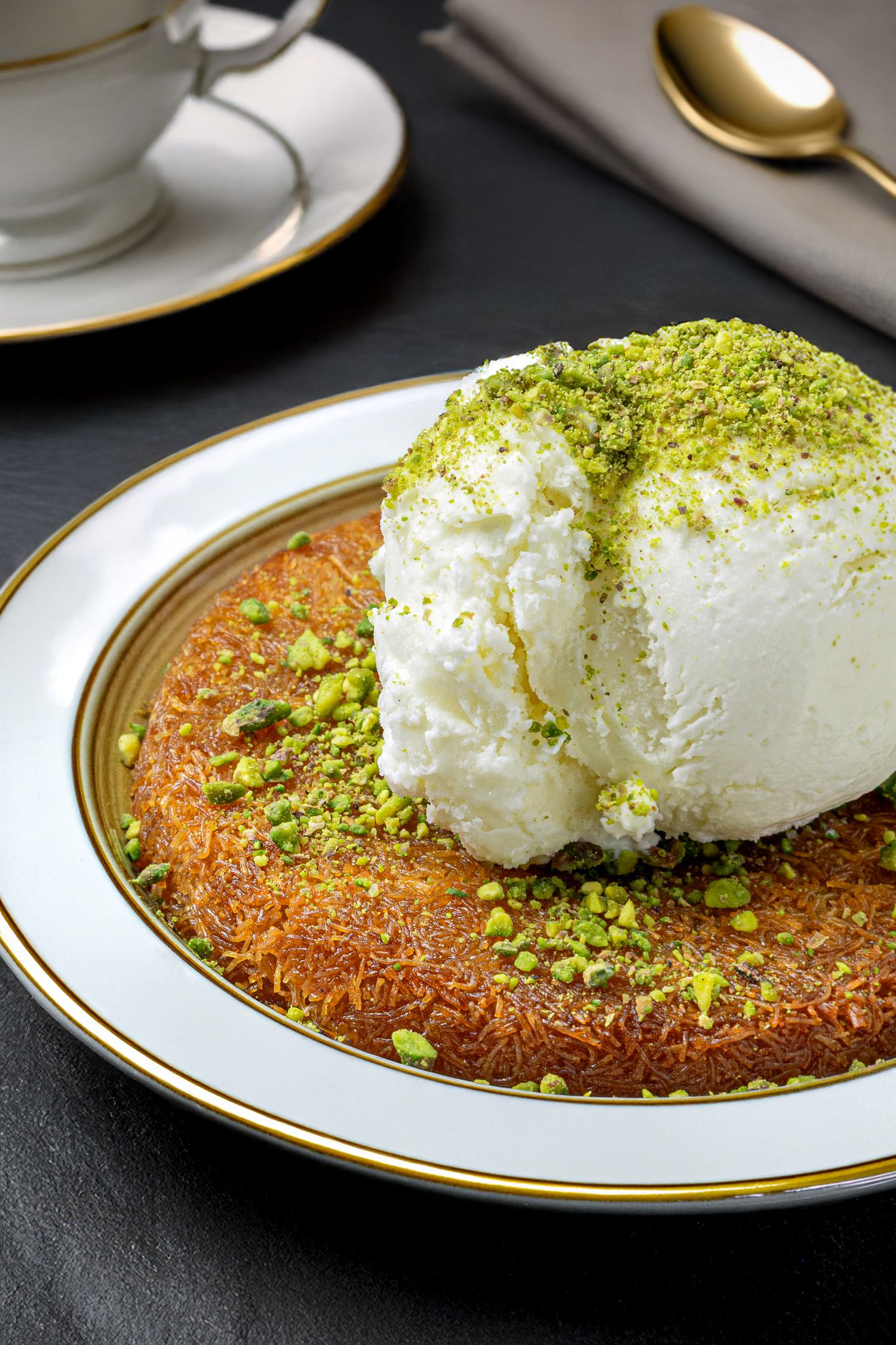 Turkish kunefe dessert topped with vanilla ice cream and crushed pistachios on an elegant gold-rimmed plate.