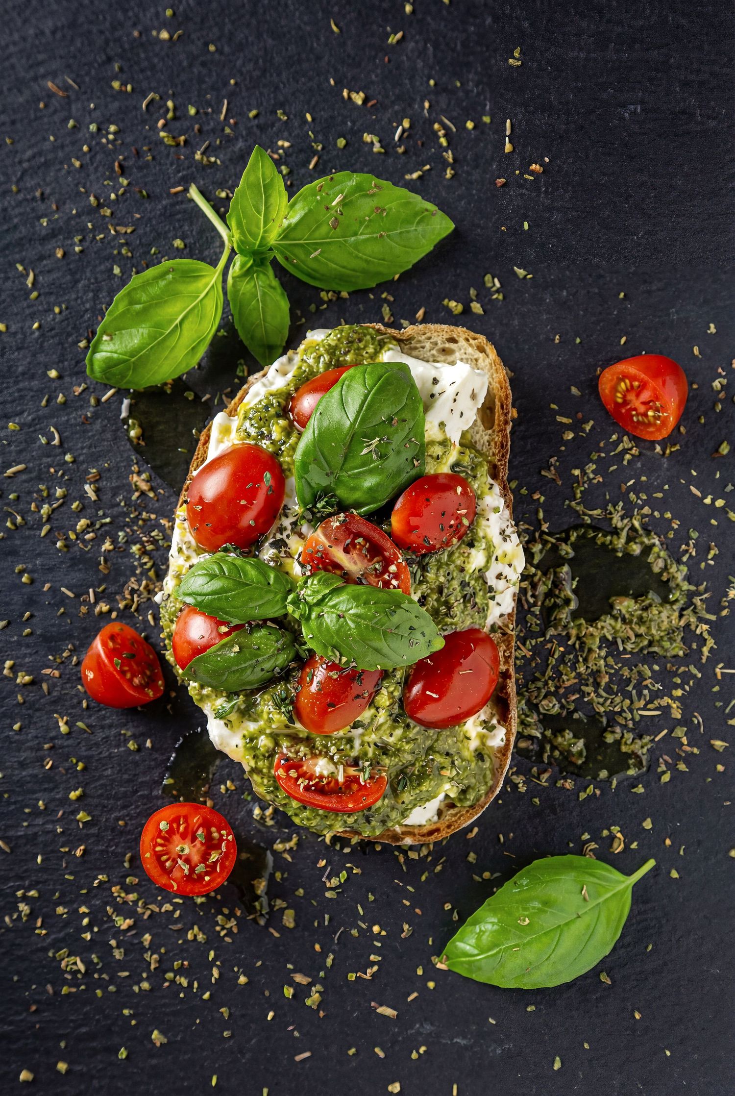 Fresh bruschetta topped with pesto, mozzarella, cherry tomatoes and basil leaves on a dark slate background.