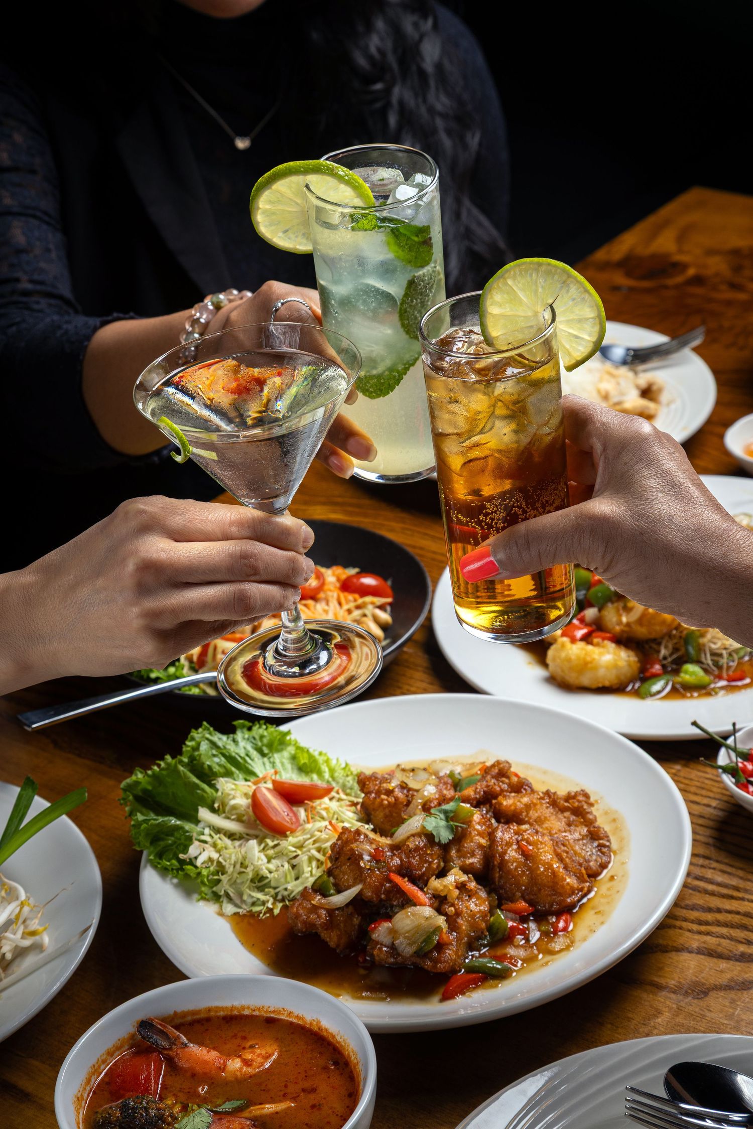 Three people toasting cocktails over a table of Thai dishes including fried chicken, curry, and fresh salad.