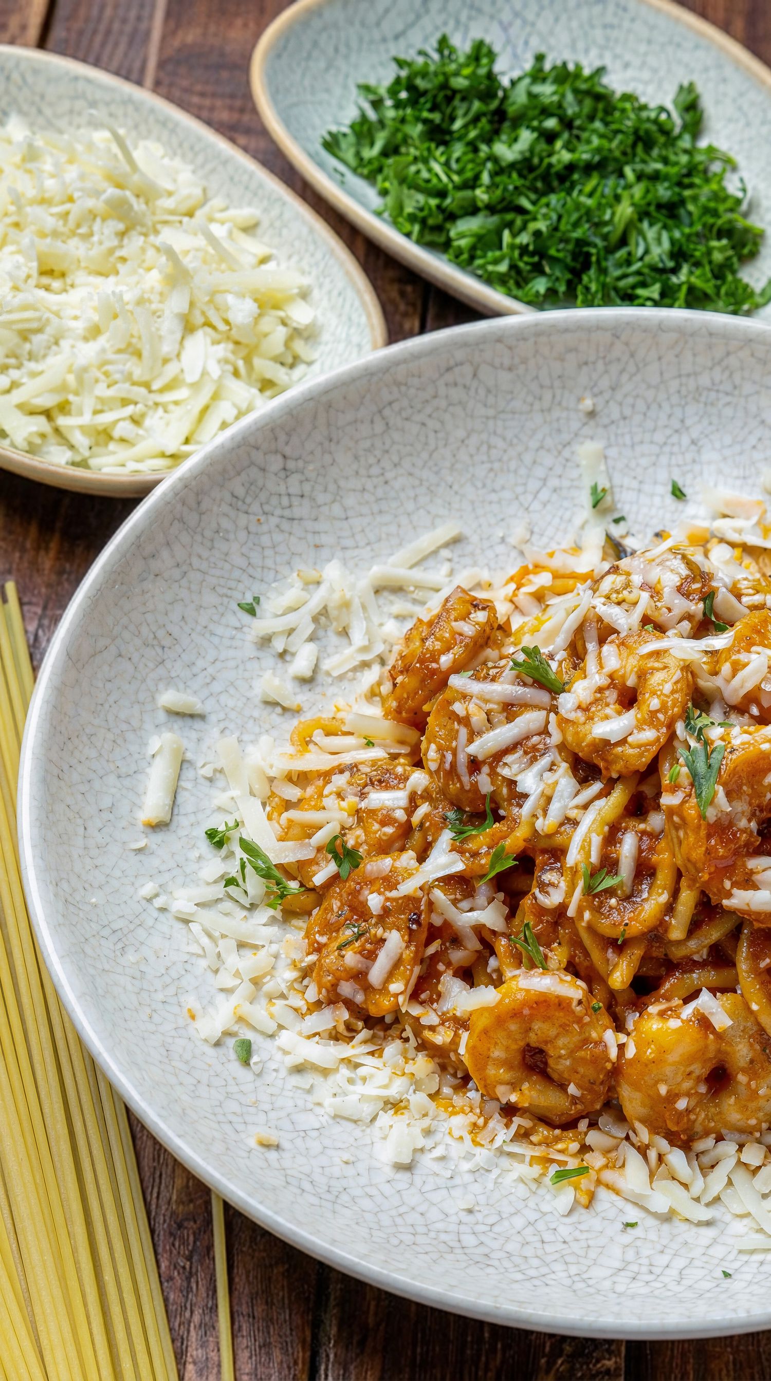 Shrimp pasta dish served with grated cheese and fresh chopped parsley on a gray ceramic plate with ingredients on side.