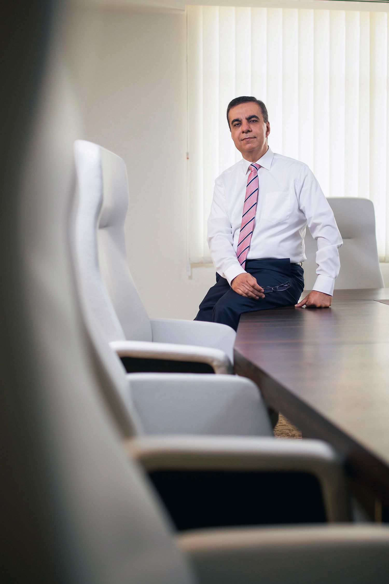 A confident businessman in a white shirt and pink tie leans against a conference table in a modern boardroom.