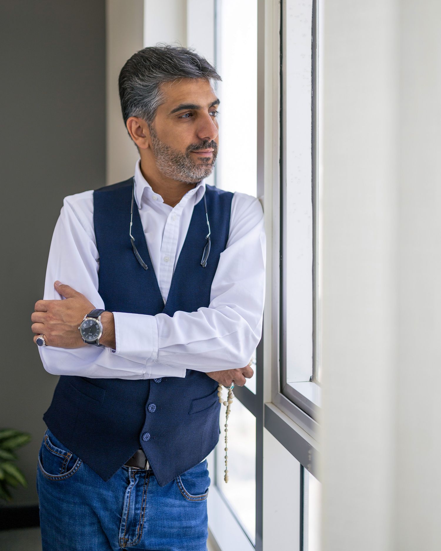 Middle-aged man with gray hair and beard in blue vest and white shirt stands arms crossed by a window.
