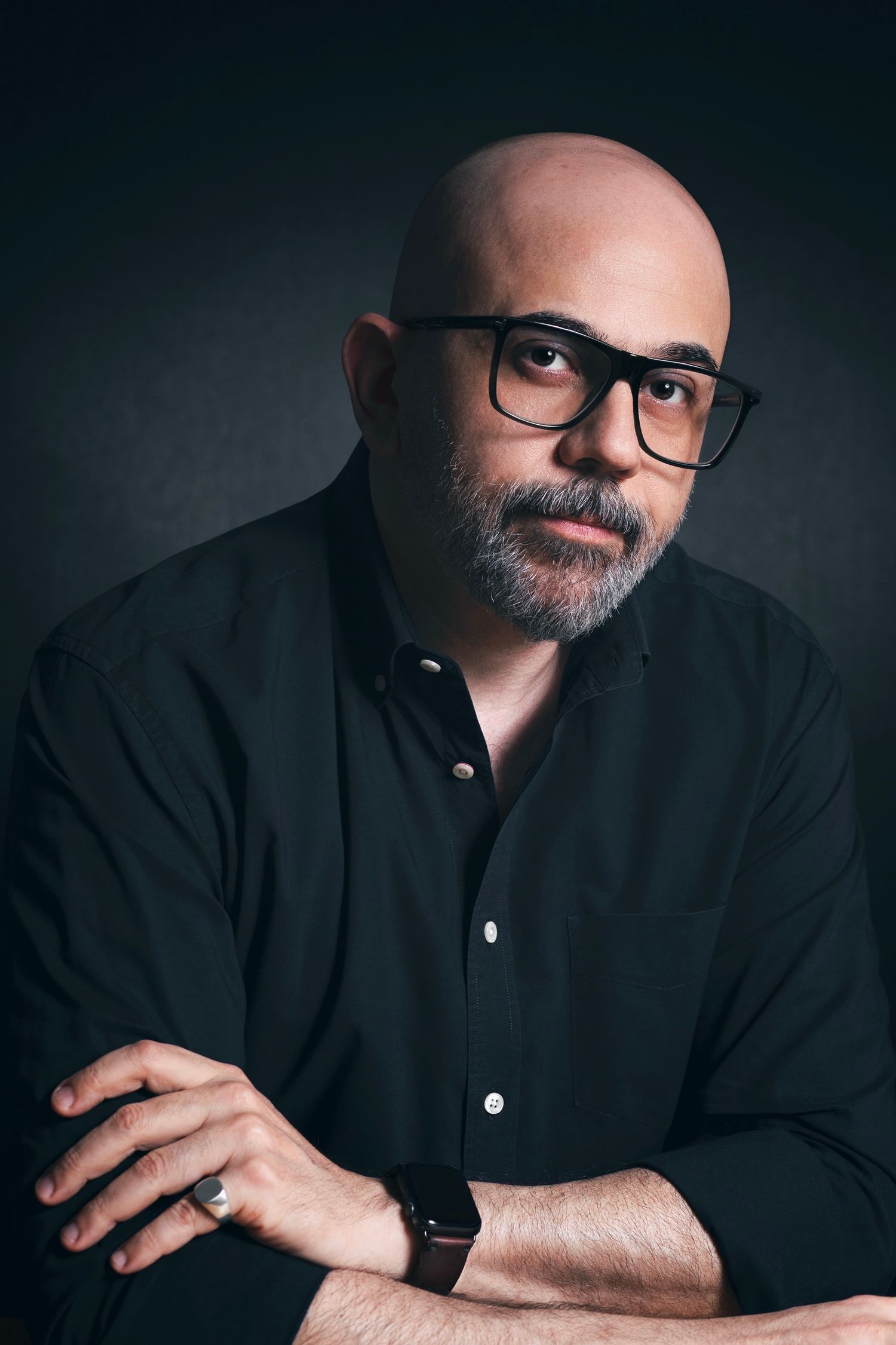Bald man with beard and glasses wearing black shirt, arms crossed, posing against dark background.