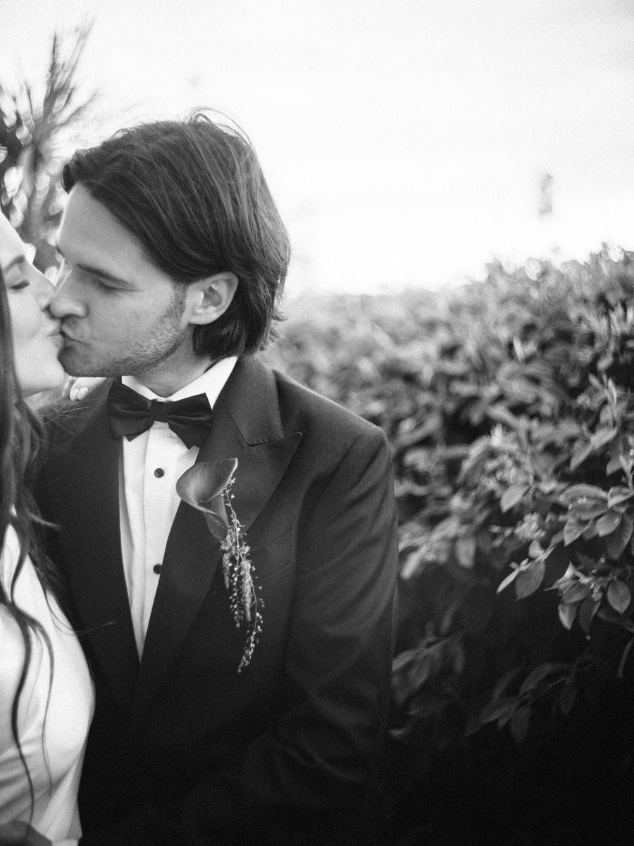 Black and white photo of a couple sharing a kiss at their wedding, surrounded by lush greenery.