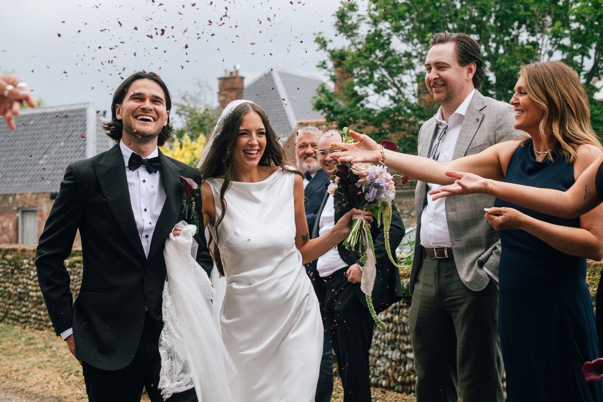 Joyful newlyweds laugh as wedding guests throw colorful confetti outside church.