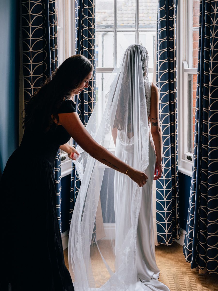 Bridesmaid in black dress adjusting bride's long lace veil by a patterned window before the wedding.