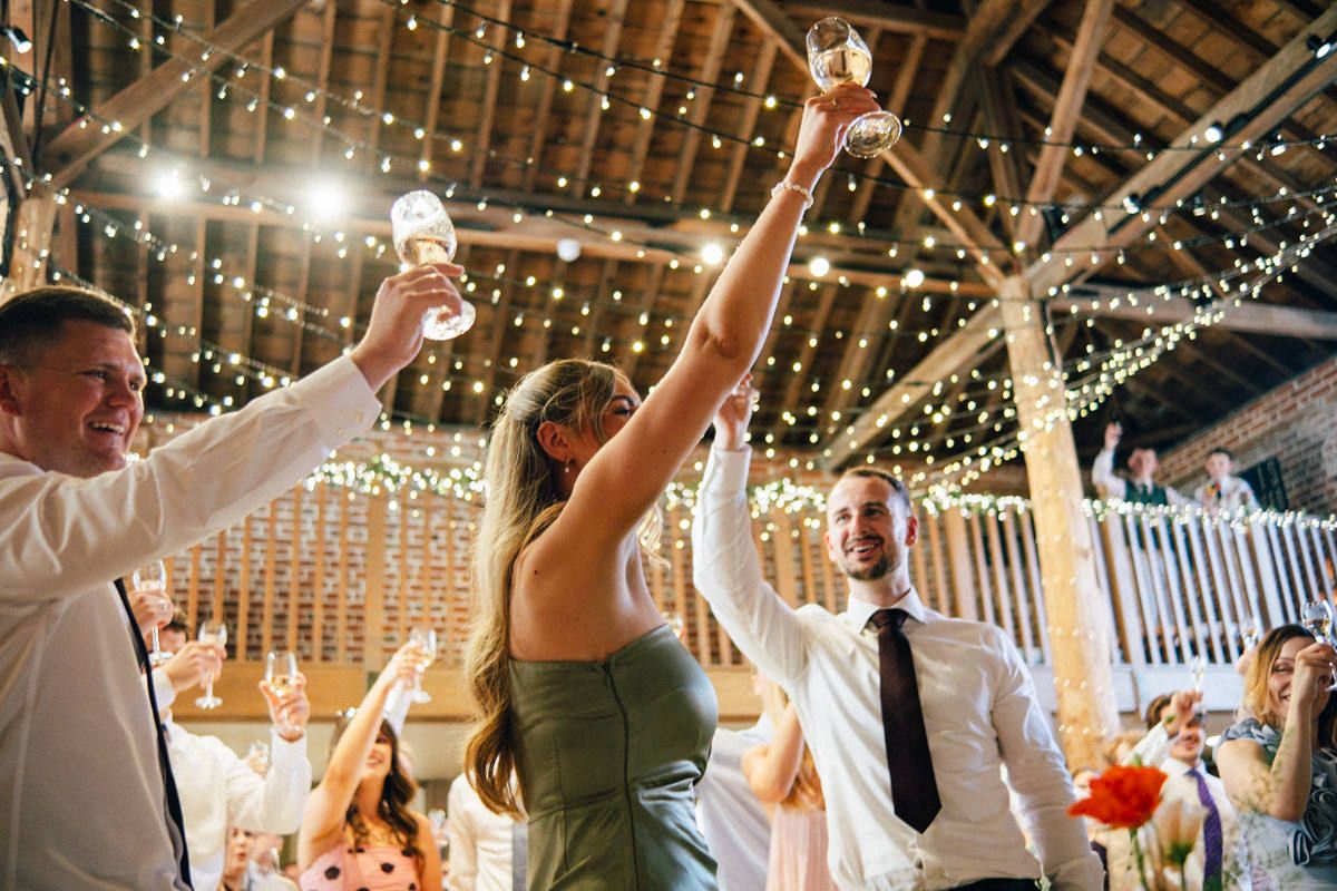 Wedding guests raise glasses in a joyful toast under fairy lights in a rustic barn venue.