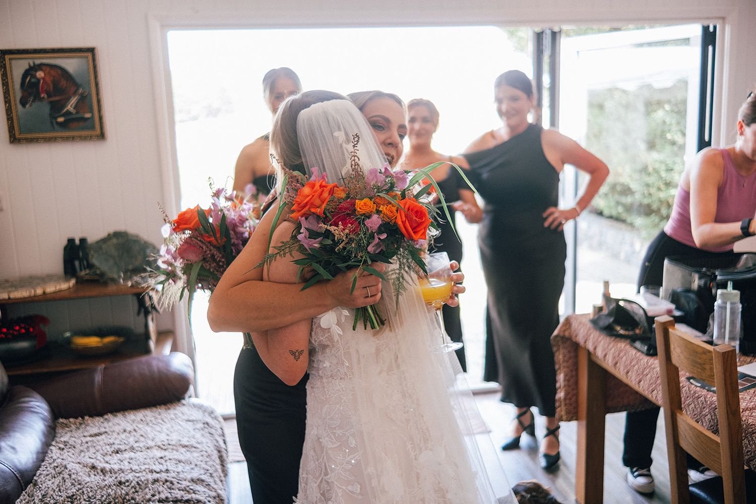 Bride receives warm hug while holding vibrant bouquet of orange and purple flowers during wedding preparations.