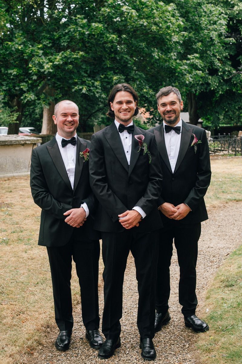 Three groomsmen in black tuxedos with bow ties and boutonnieres posing outdoors at a wedding.