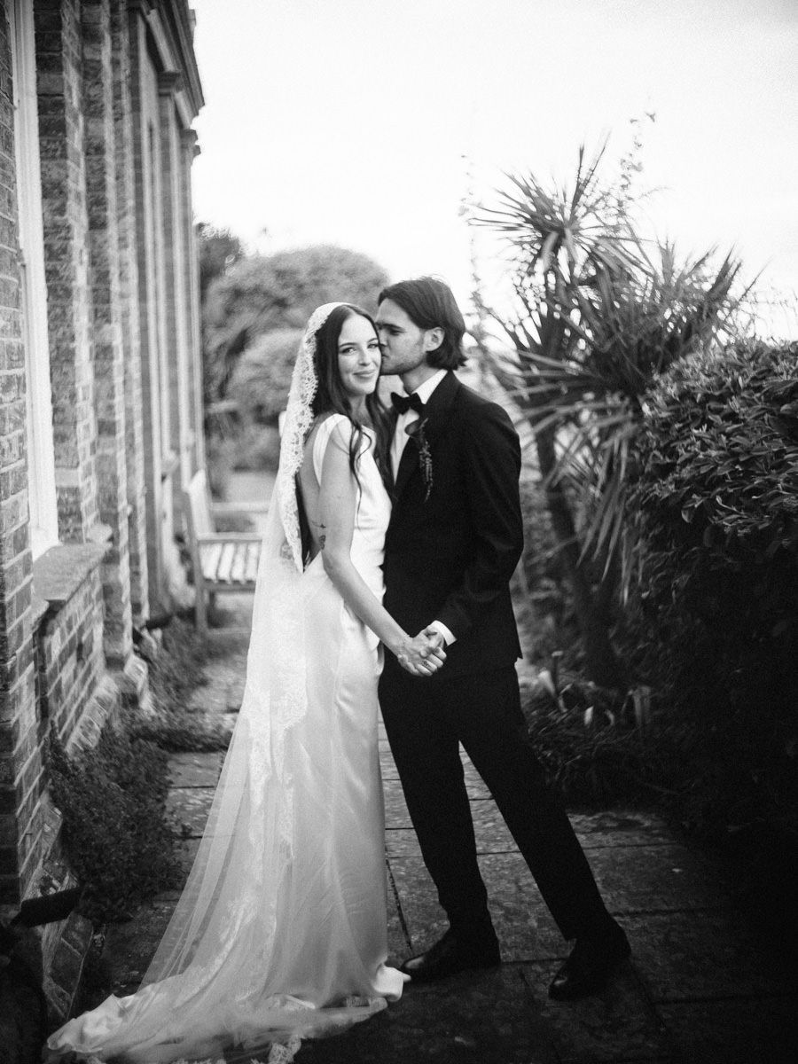 Black and white wedding portrait of a bride in a flowing gown and groom holding hands beside a brick building.