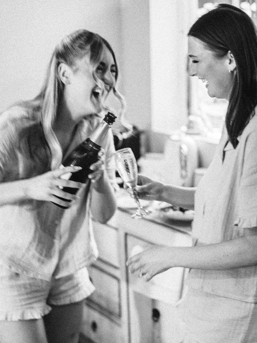 Two women laughing while pouring champagne in black and white wedding preparation photo.
