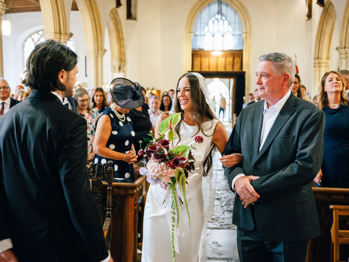 Bride smiling at groom as father walks her down the aisle in a church.