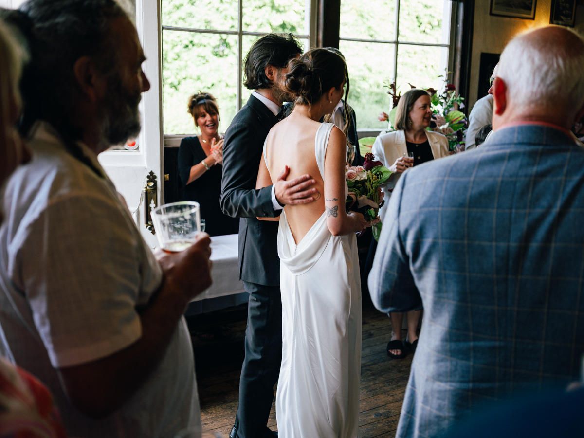 Wedding guests mingle indoors as a bride in a backless white dress holds a bouquet with her groom.