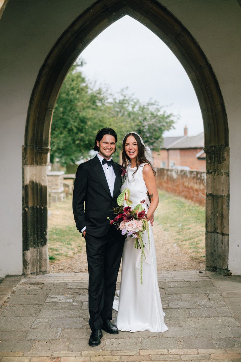 Bride and groom posing under a stone Gothic arch at a church, bride holding a colorful bouquet.