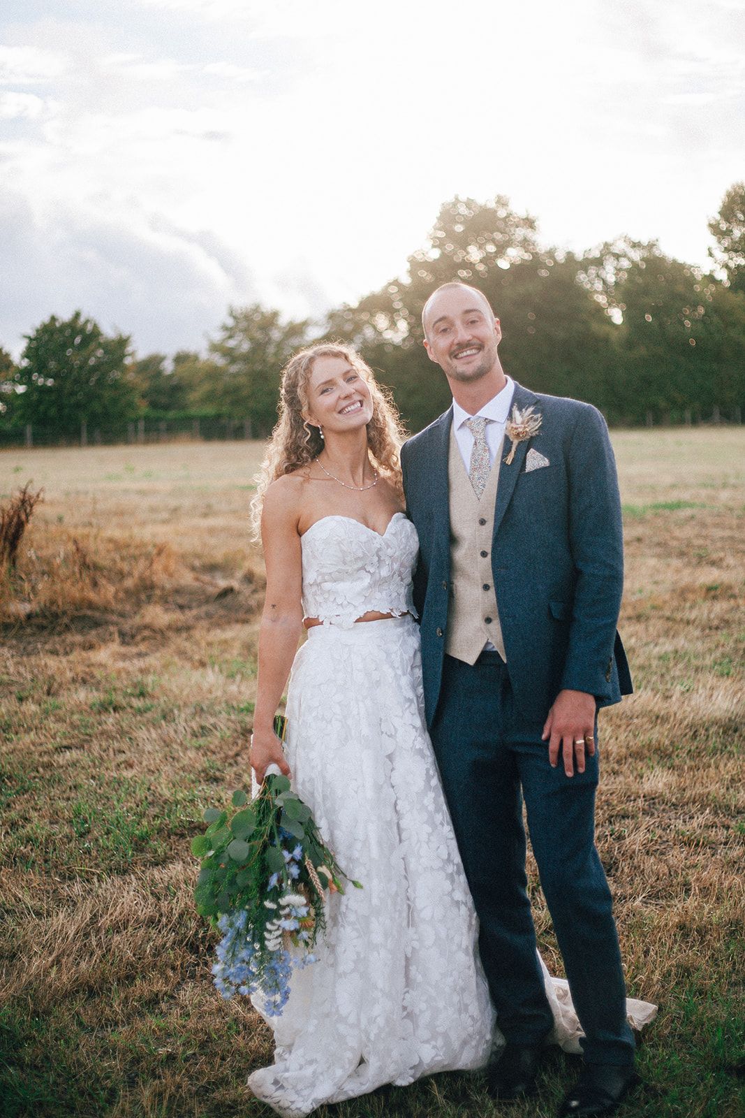 Smiling bride and groom posing in a golden field at sunset, bride holding a blue and green bouquet.
