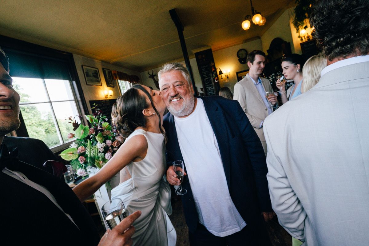 A cheerful older man in a navy blazer receives a kiss on the cheek at a lively wedding reception.