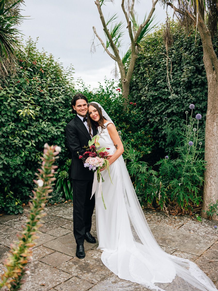 Bride and groom embracing in a lush garden, bride wearing a long veil and holding a bouquet.