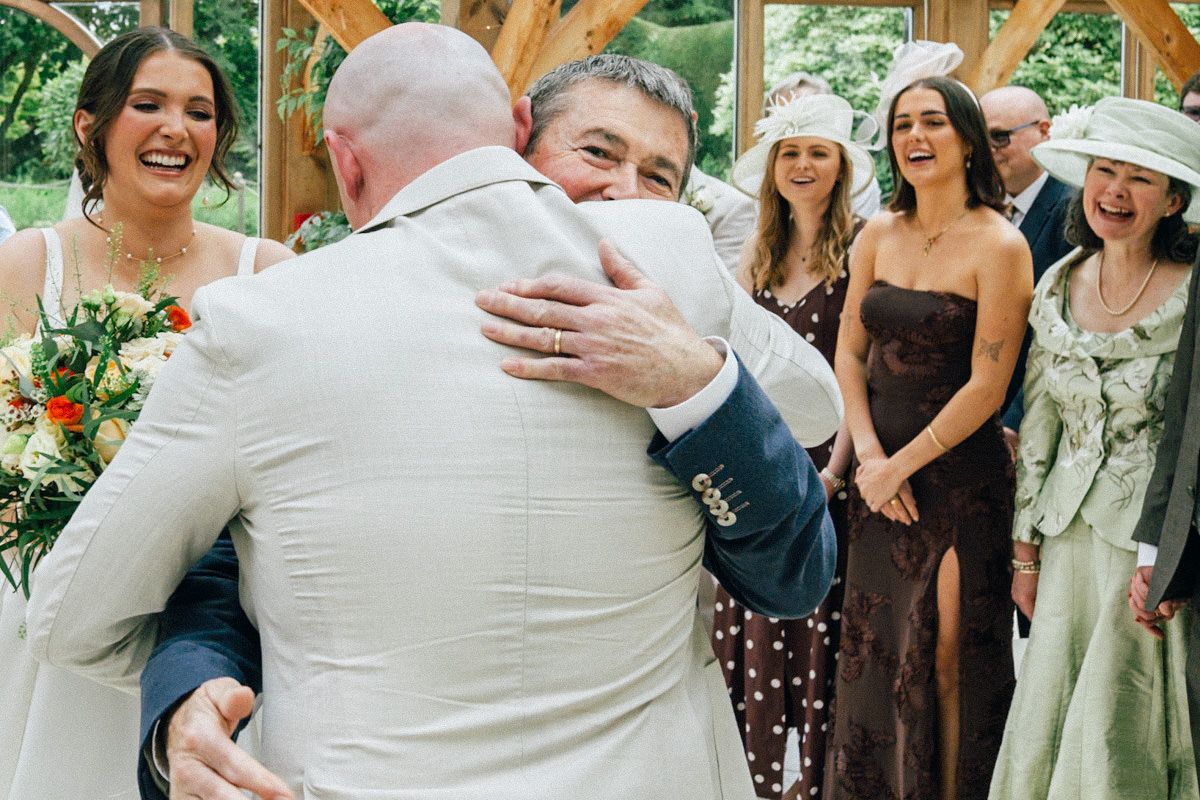 Groom sharing emotional hug with guest while bride laughs during wedding reception.