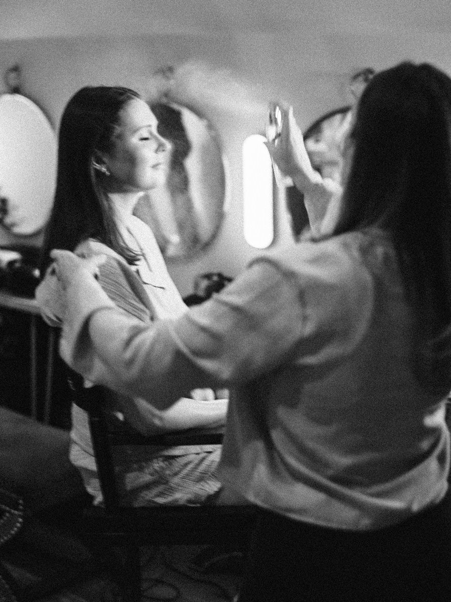 Black and white photo of bride getting makeup applied at vanity mirror before wedding.