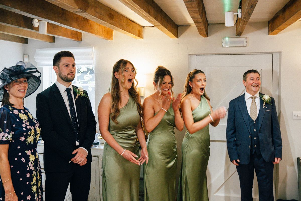 Wedding party reacting with joy as bridesmaids in sage green dresses see the bride.
