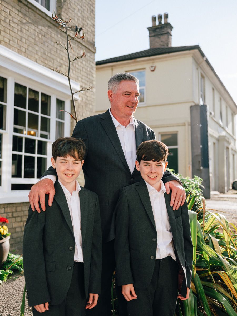 A tall man in a dark suit poses proudly with two young boys in matching suits outside a venue.