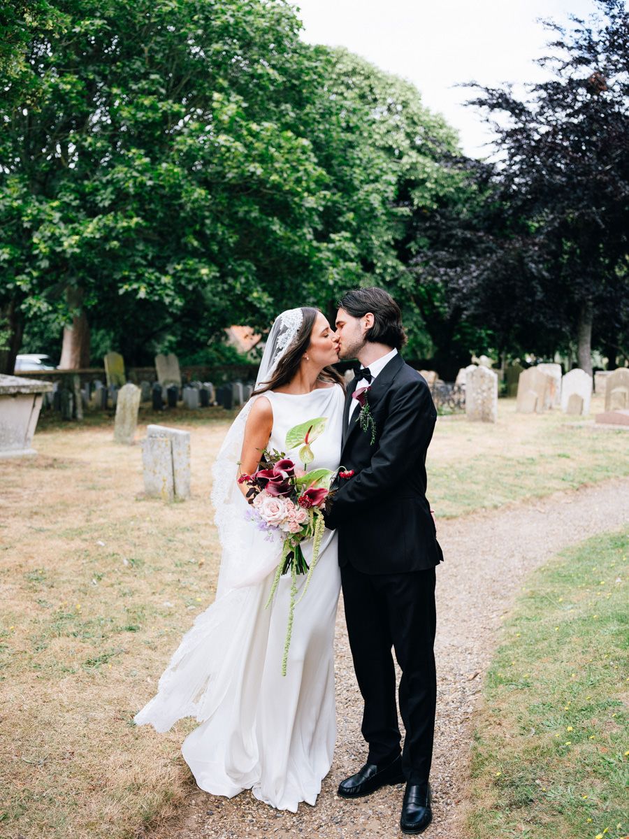 Bride and groom sharing a kiss in a churchyard, bride holding a lush burgundy and pink bouquet.