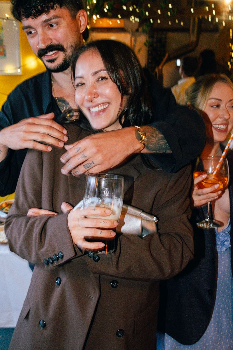 Woman in brown blazer smiles while being hugged from behind at a warmly lit evening celebration.