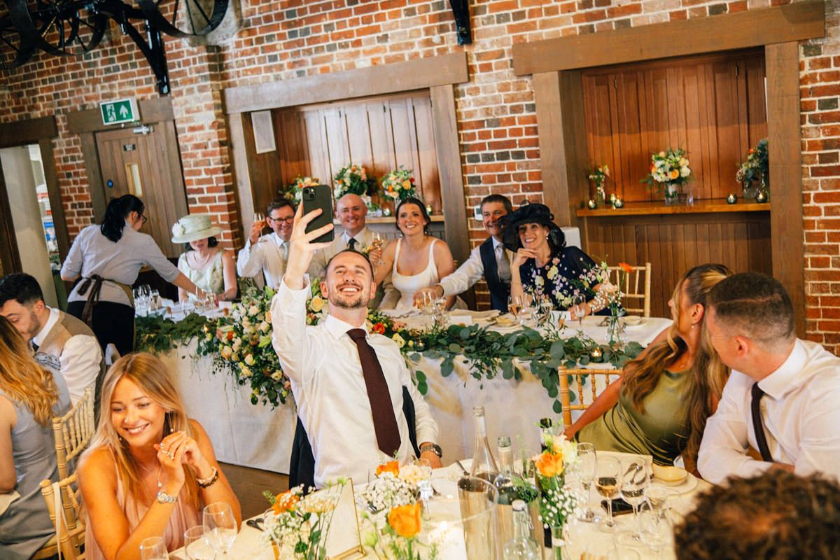Groom takes a selfie with laughing wedding guests at a beautifully decorated reception table.