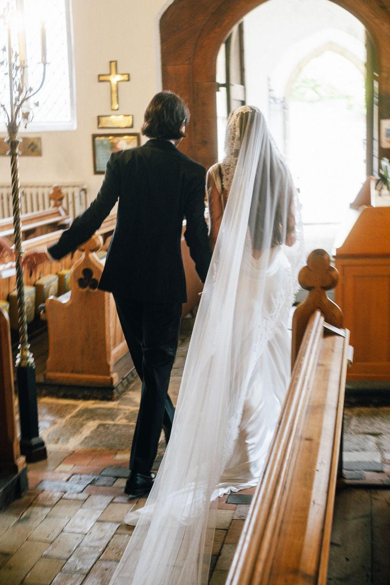 Bride in cathedral veil walks down church aisle with escort past wooden pews.
