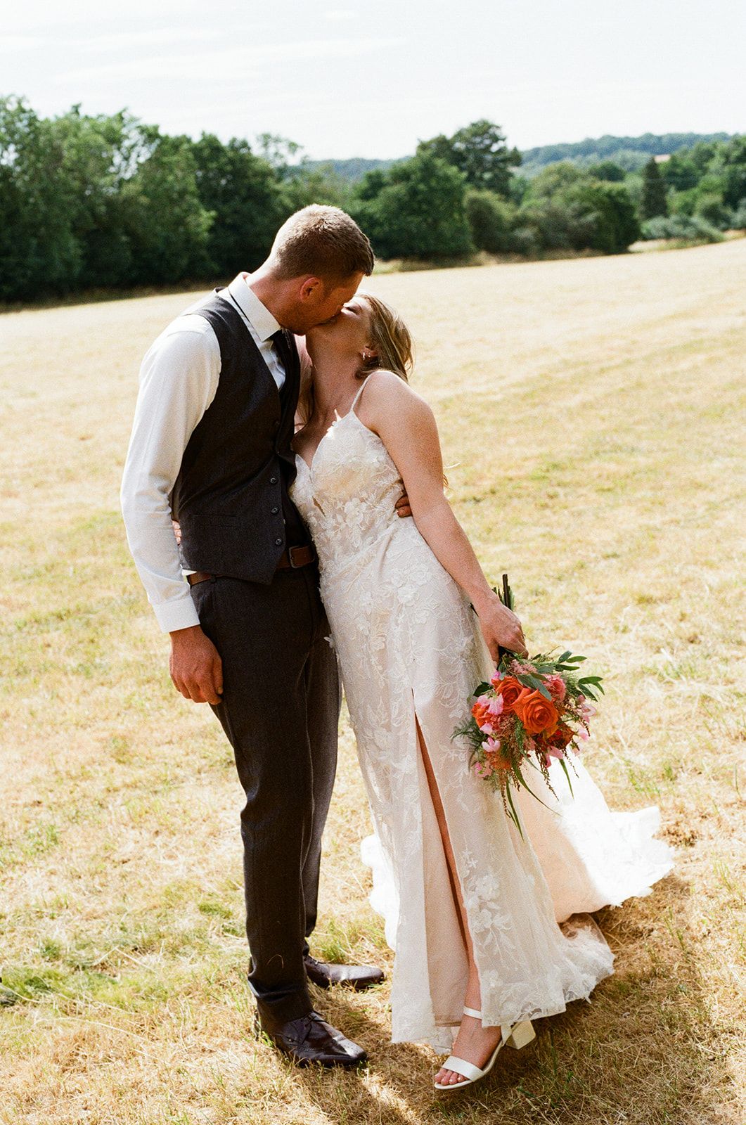 Bride and groom share romantic kiss in sunny field holding orange bouquet.