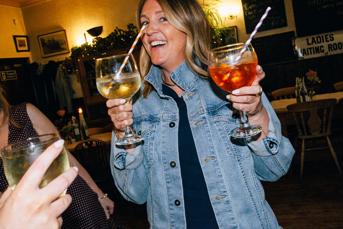A smiling woman holds two cocktails at a lively wedding reception bar.