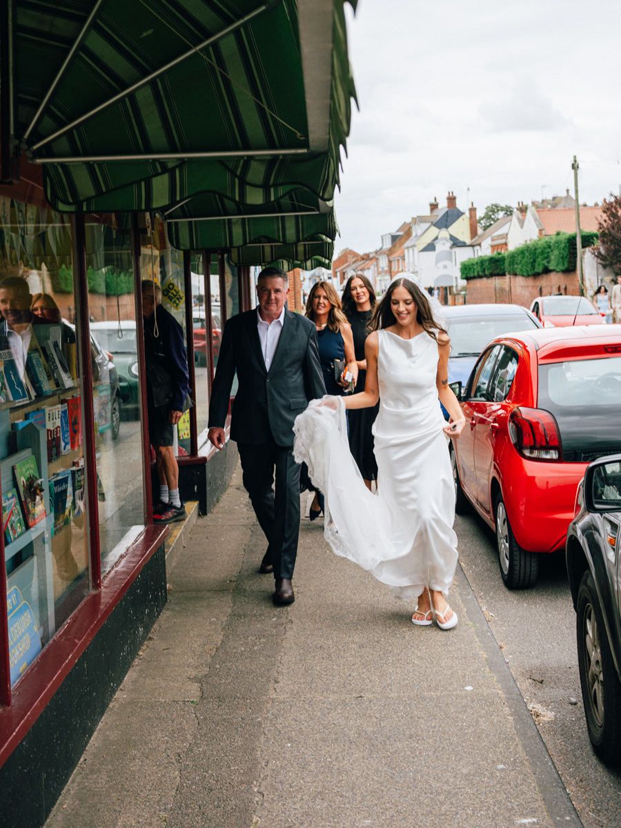 Bride in elegant white gown walking along a town street with wedding party before the ceremony.