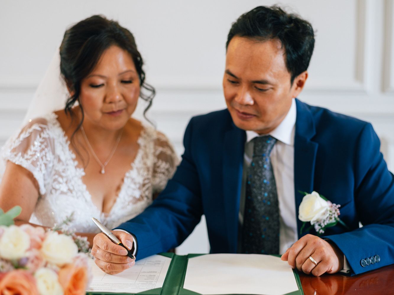Bride and groom signing wedding certificate at elegant ceremony, bride in lace dress, groom in blue suit with boutonniere.