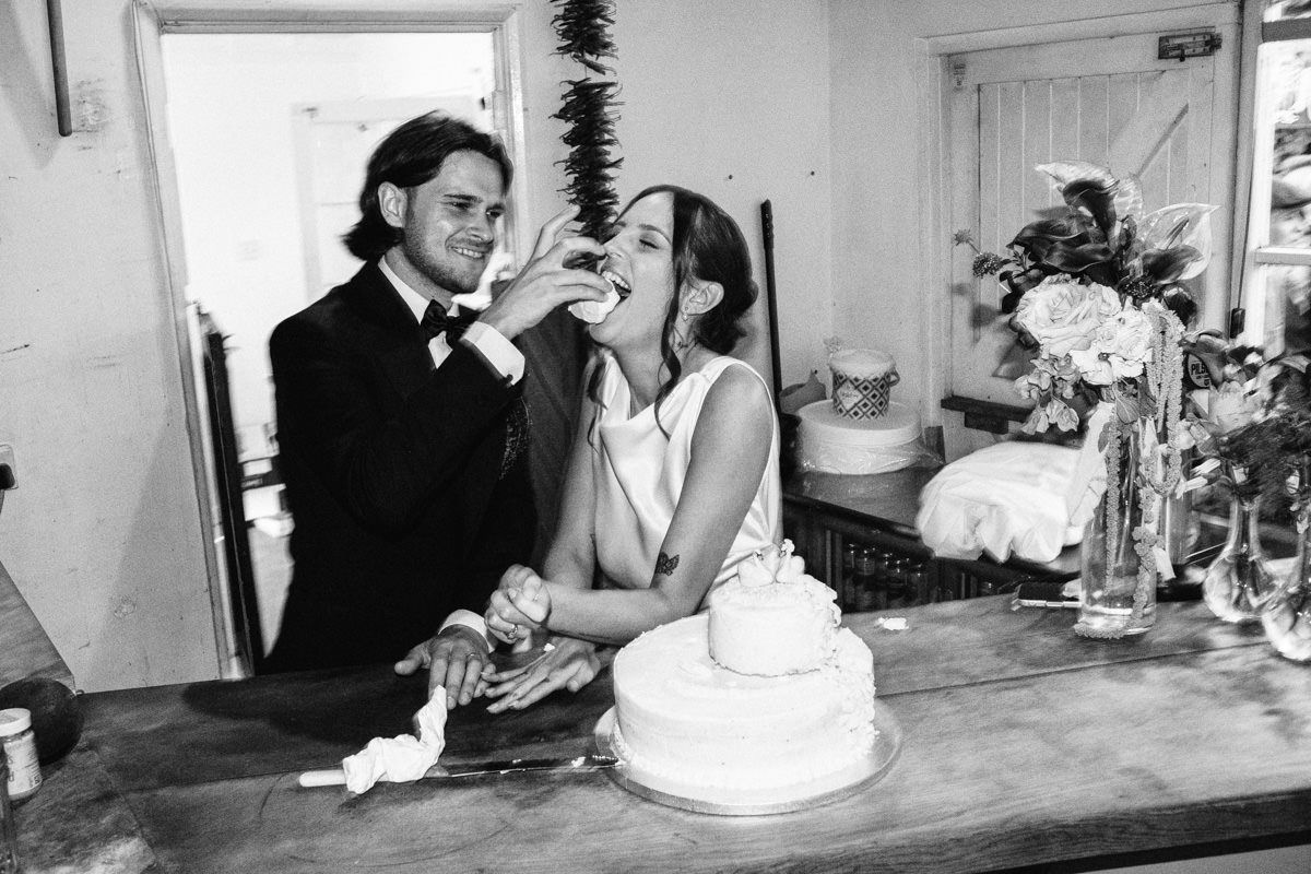 Black and white photo of groom feeding bride wedding cake as they share a joyful moment together.