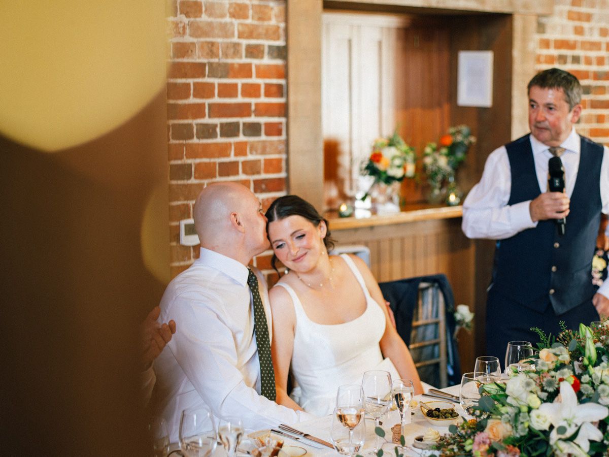 Groom tenderly kisses bride during heartfelt wedding speech in a charming rustic brick venue.