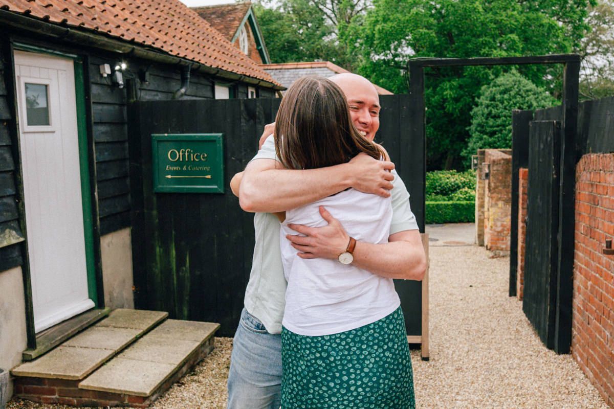 Two people sharing a warm embrace outside a rustic office building on a sunny day.