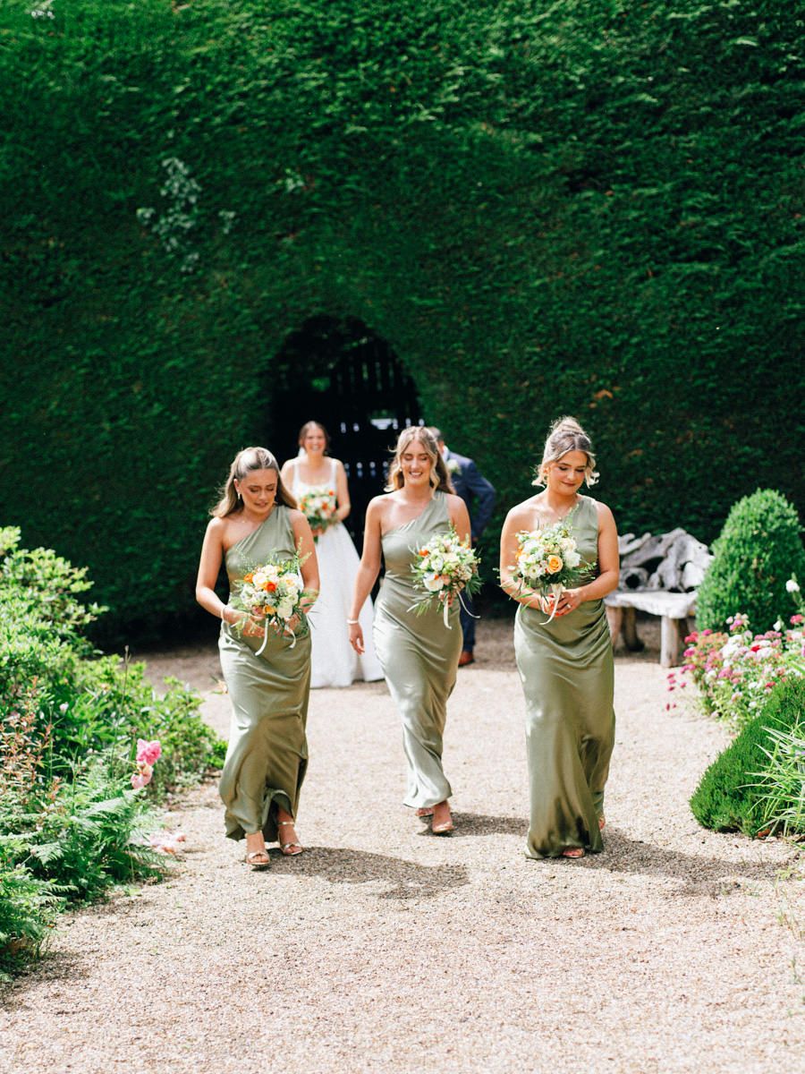 Three bridesmaids in sage green gowns holding bouquets walking through garden pathway.