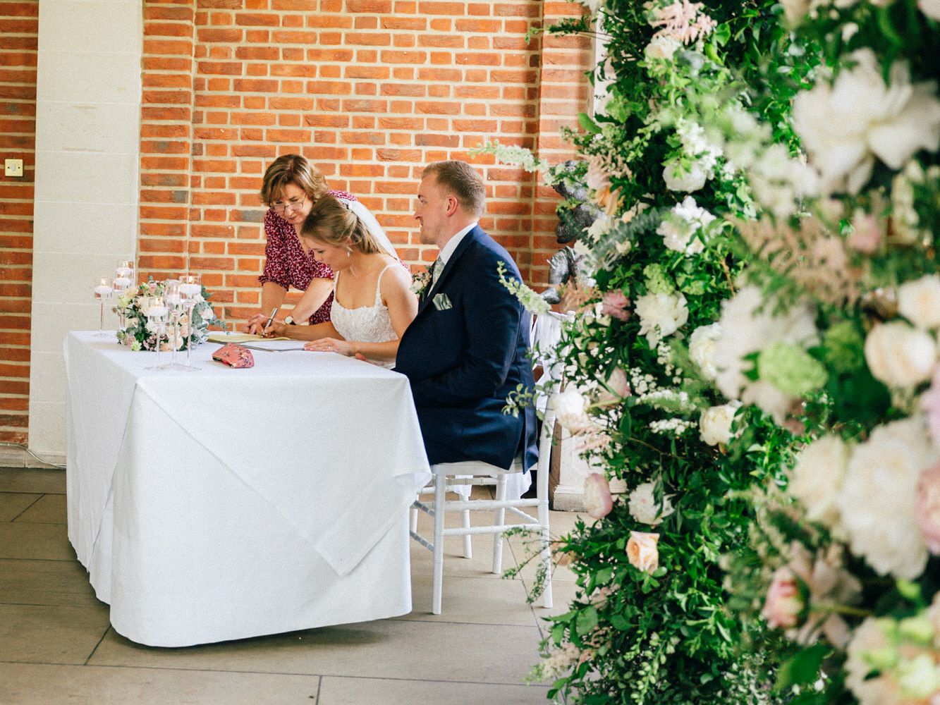 Bride and groom signing wedding register at white table with floral wall backdrop and brick interior.
