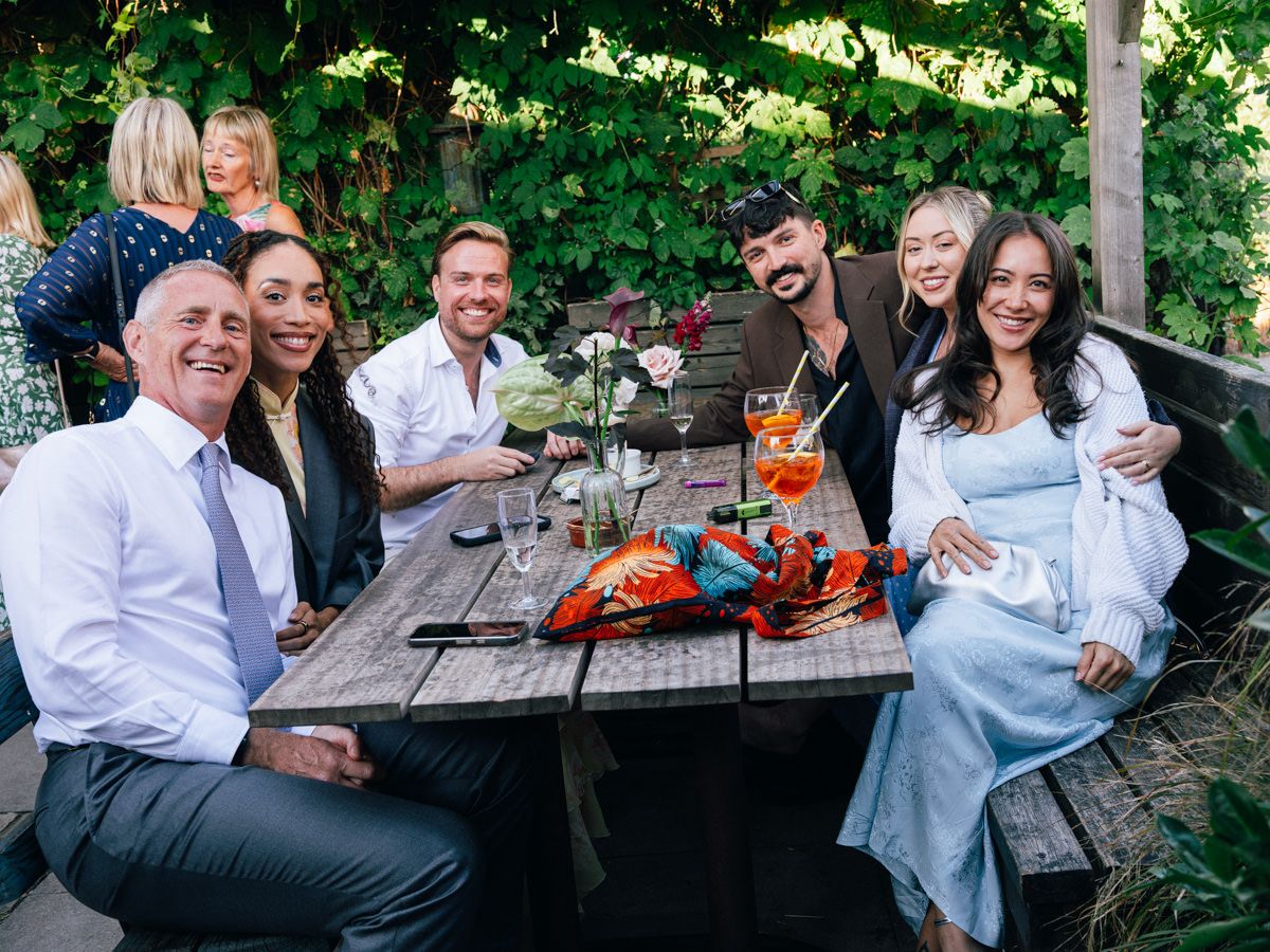 A joyful group of six wedding guests smile and pose together at an outdoor dining table with lobster.