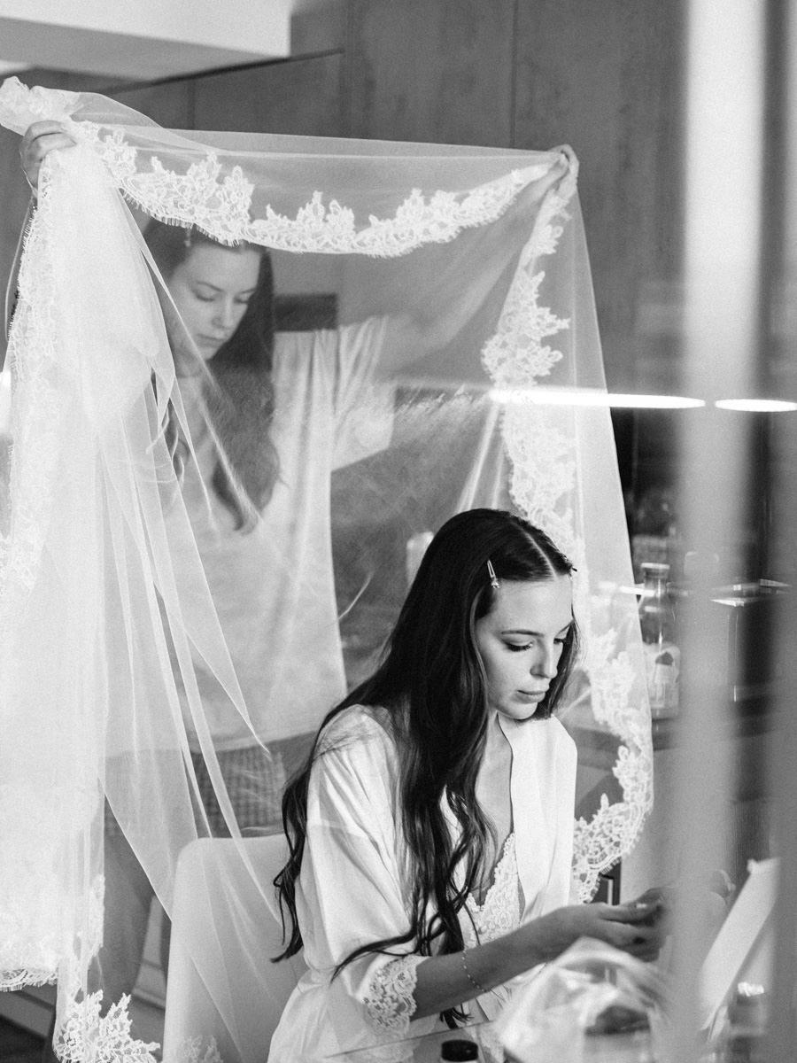 Black and white photo of a bride getting ready with her lace veil being held up behind her.