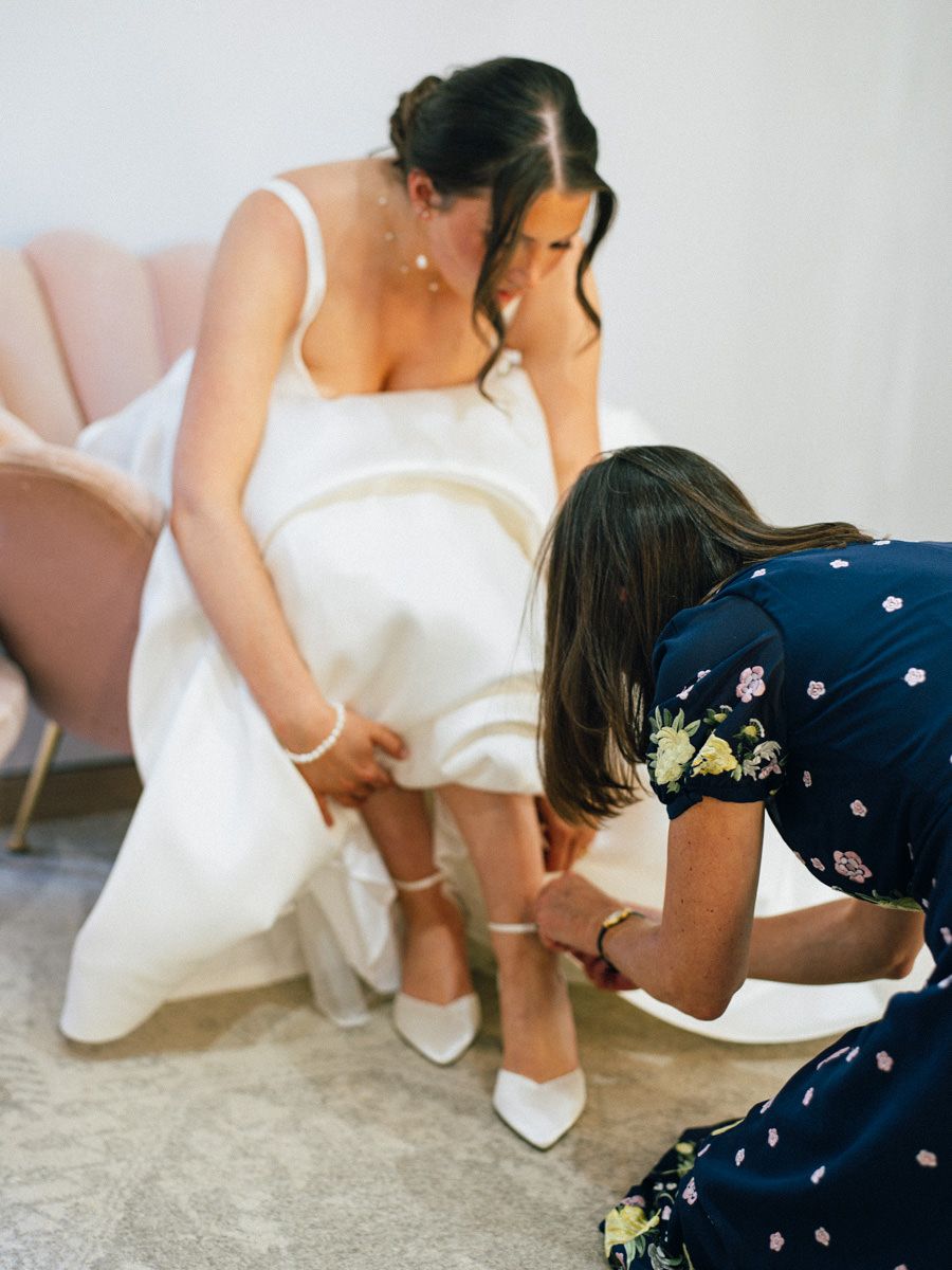 Bridesmaid in navy dress helping bride put on white heels before the wedding ceremony.