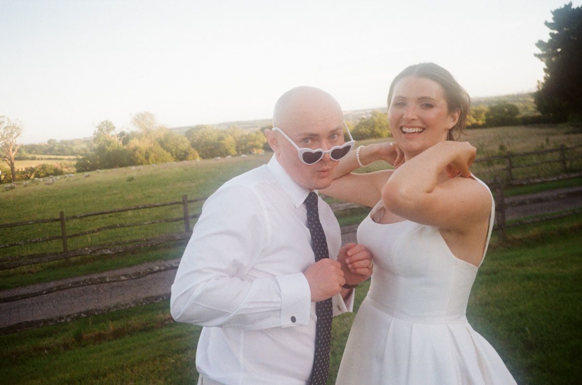 A bride and guest playfully pose with white sunglasses outdoors at a sunny wedding venue.