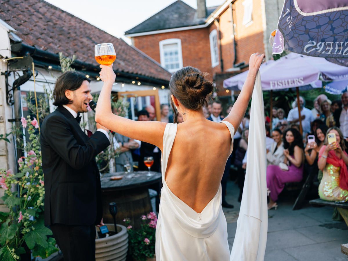 A bride in a backless dress and groom raise drinks triumphantly before cheering wedding guests outdoors.